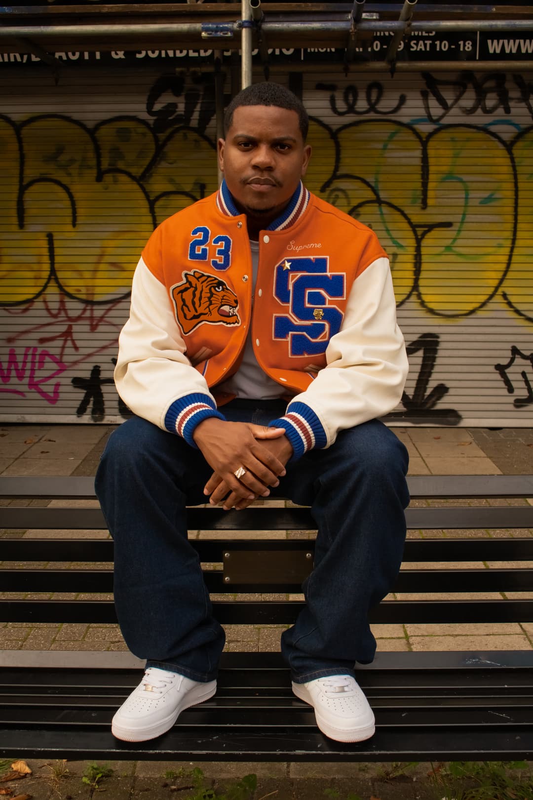 Black man sat on a bench wearing orange jacket blue trousers and white shoes