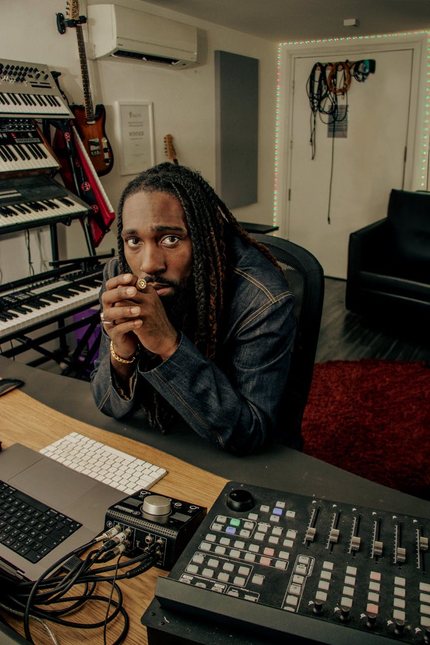 A black man sat in front of a desk with music studio equipment played on the desk